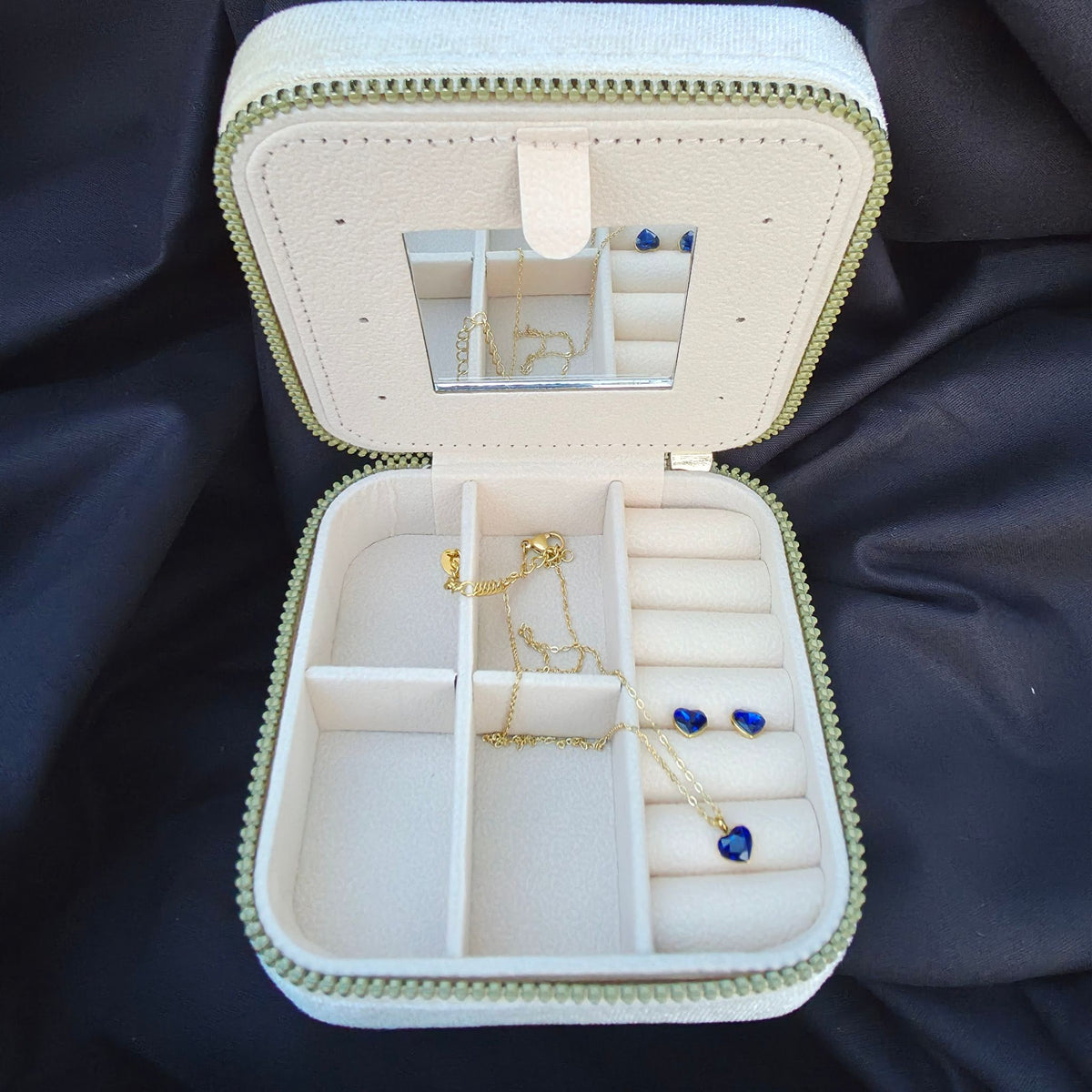 Avattu beige samettinen matkakorurasia, jonka sisällä on kullanvärinen Bohm Paris -sydänkaulakoru ja yhteensopivat sydänkorvakorut safiirinsinisillä Swarovski-kristalleilla. Kannessa sisäänrakennettu peili.